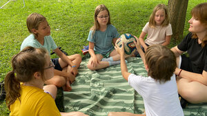 Bild zeigt Kinder bei der Freizeit 5 days, die auf einer Picknickdecke im Kreis sitzen und einen Ball herumwerfen.